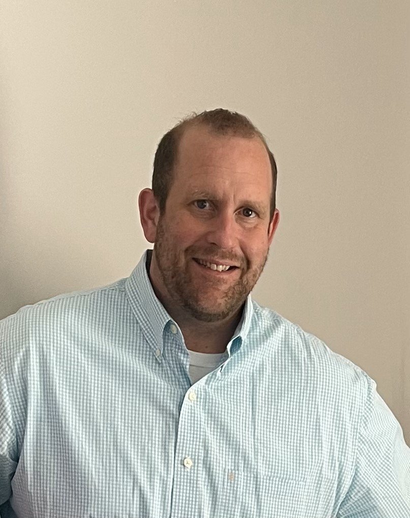 A man with short hair and a beard smiling, wearing a light blue checkered button-up shirt, standing in front of a plain beige wall.