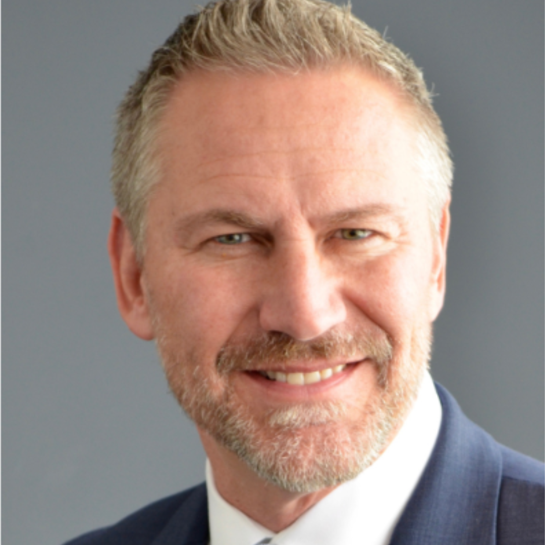 A smiling middle-aged man with a beard and short blond hair, wearing a suit and tie, against a gray background.