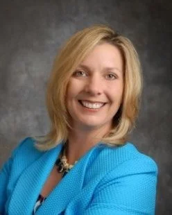 Portrait of a woman with shoulder-length blonde hair, smiling, wearing a bright blue blazer and a black necklace, against a dark background.