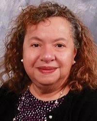 A woman with curly hair and a slight smile, wearing a dark top with white dots, posing against a gray background.