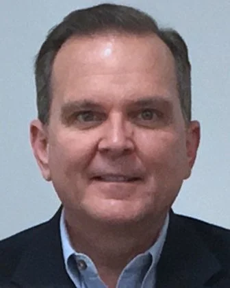 Close-up photograph of a middle-aged man with short brown hair, wearing a dark blazer and light blue collared shirt, smiling softly against a plain light-colored background.