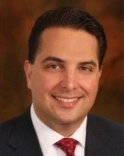Professional portrait of a smiling man with dark hair, wearing a suit and red tie.