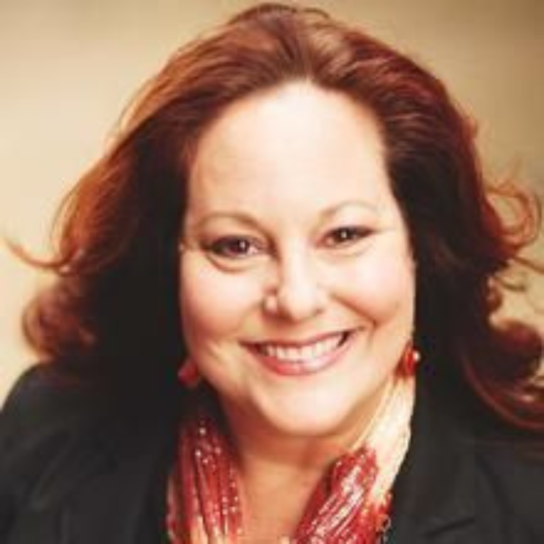 A woman with red hair, smiling, wearing a black top and red patterned scarf and earrings.