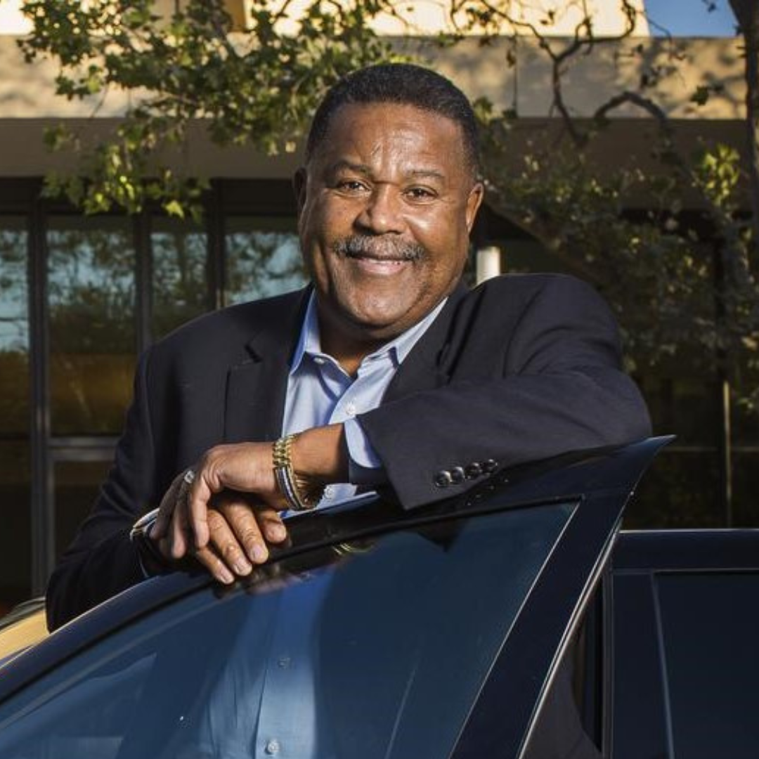 A smiling man in a dark suit leaning on a car with a building and trees in the background.
