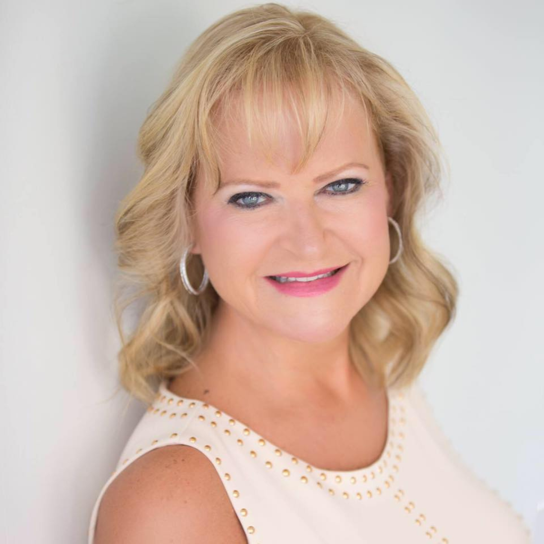 A woman with blonde hair, blue eyes, and hoop earrings smiling at the camera, wearing a sleeveless white top with gold studs, against a plain white background.
