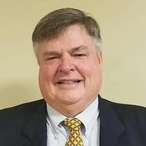 A middle-aged man with gray hair, wearing a black suit, white shirt, and yellow patterned tie, smiling in front of a plain beige background.