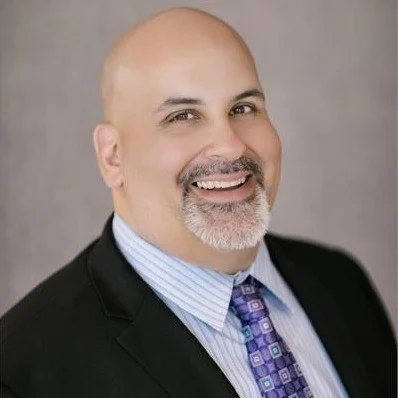 Professional headshot of a smiling bald man with a beard, wearing a black suit, light blue shirt, and purple checkered tie, against a gray background.