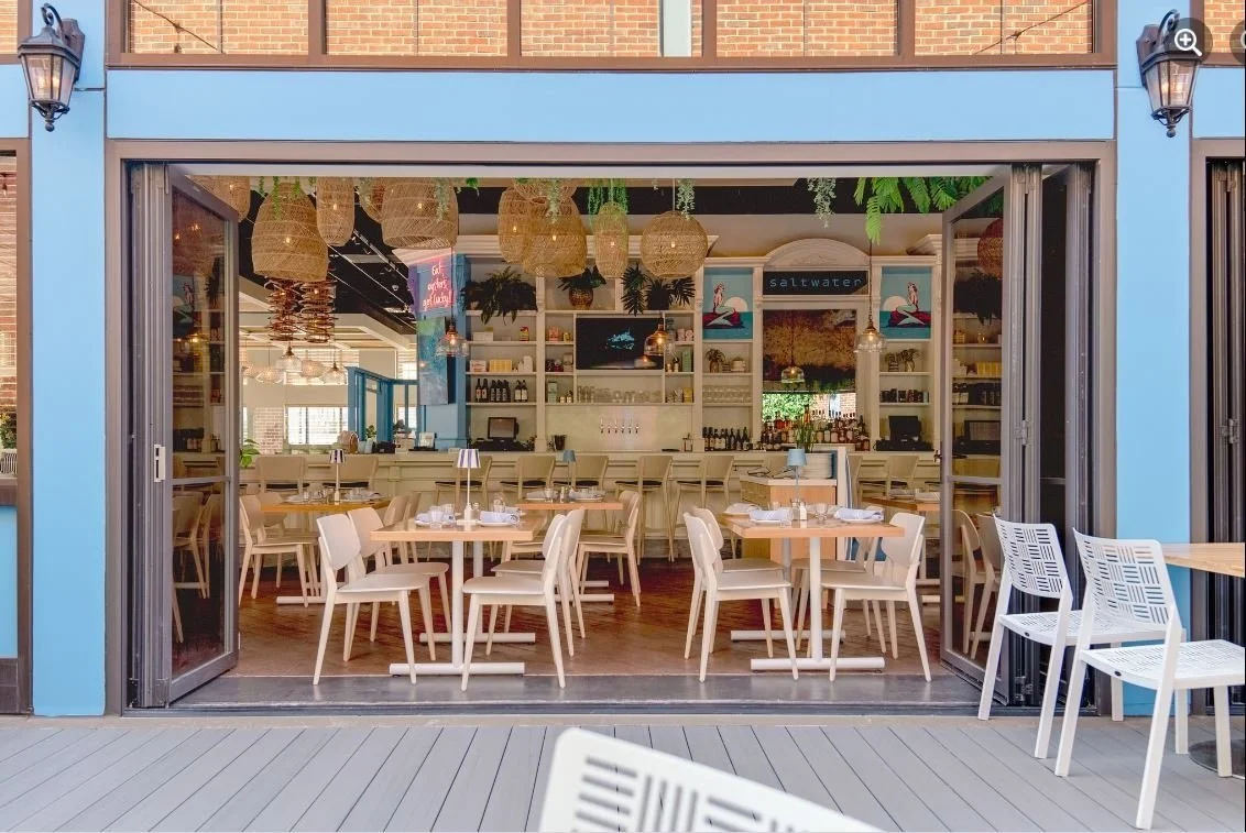 The image shows the open entrance of a modern, stylish restaurant with white chairs and wooden tables set for dining. Inside, there are hanging wicker light fixtures, shelves with bottles and decor, and a colorful mural behind the bar area. Outdoor seating is available with white chairs around tables on a gray wooden deck.