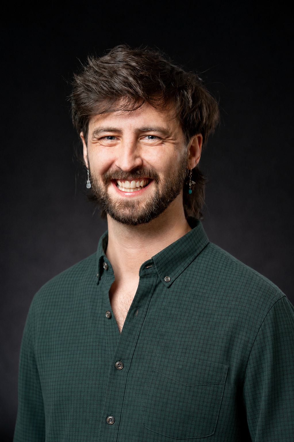 A smiling man with brown hair, a beard, and blue eyes wearing a green checkered shirt against a dark background.
