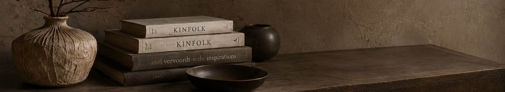 A rustic wooden table with a large textured ceramic vase, a stack of four books, a small black vase, and a black bowl, against a textured wall.