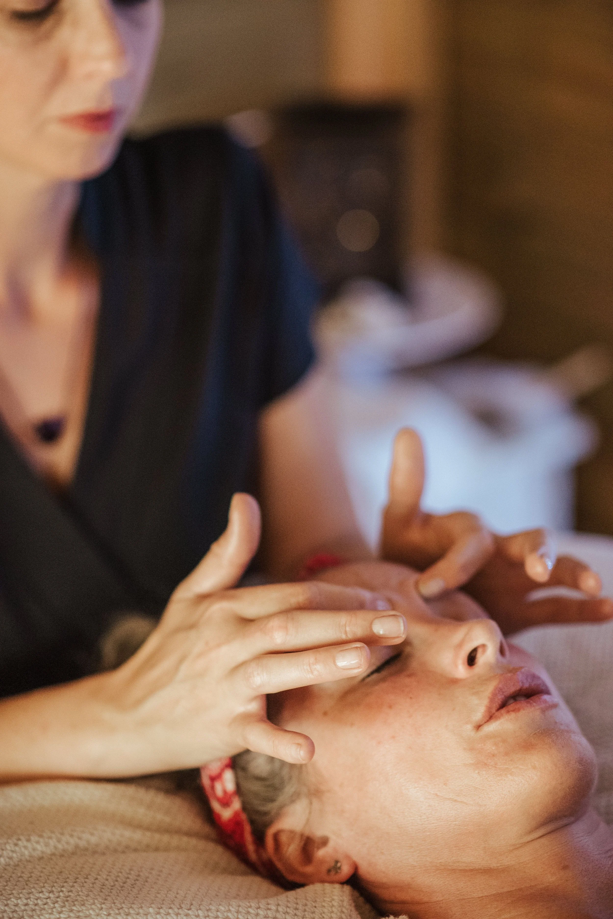 A person with short gray hair receives a facial massage from another person with nail polish on their nails.