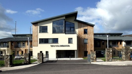 A modern residential building with a unique angled roof, large windows, and a combination of wood and white exterior walls, surrounded by a stone entrance gate.