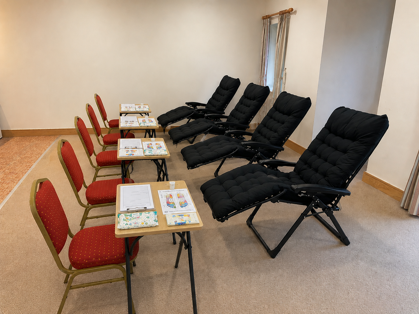 Four red chairs and four tables with papers and clipboards on a beige carpeted floor. Four black reclining chairs arranged in a row along the wall in a room with cream walls and a window with curtains.