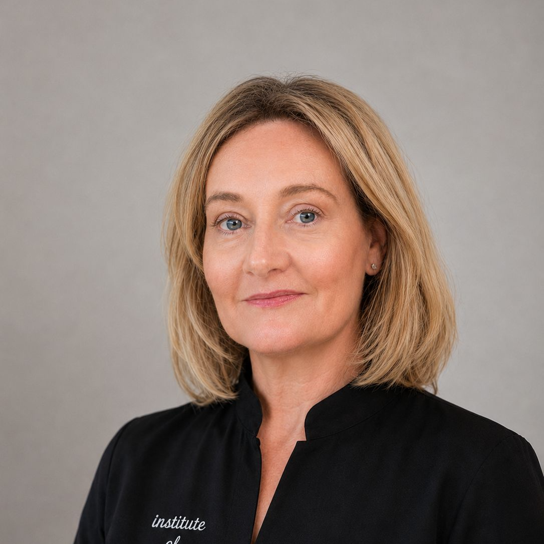 A professional woman with blonde hair wearing a black top with white embroidery, standing against a gray background.