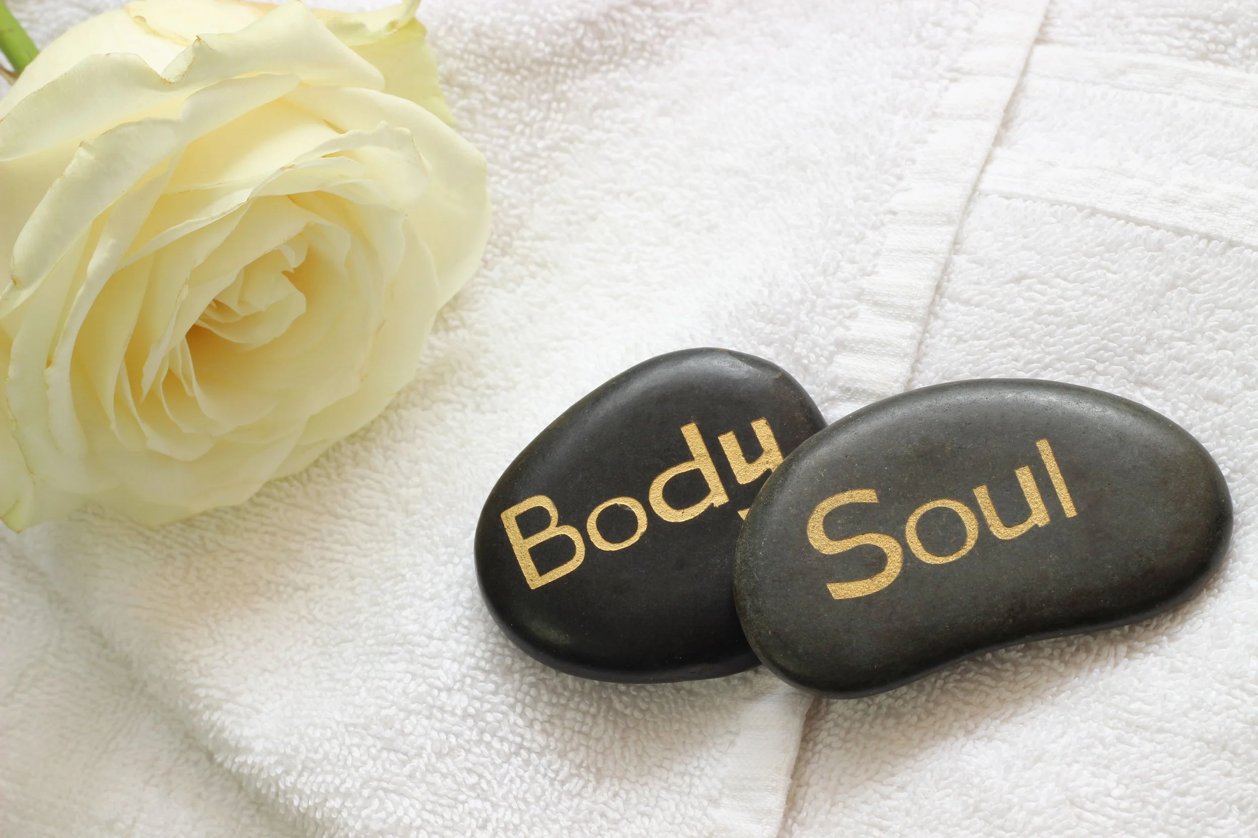 Close-up of two black stones with the words "Body" and "Soul" written in gold, placed on a white towel with a cream-colored rose beside them.