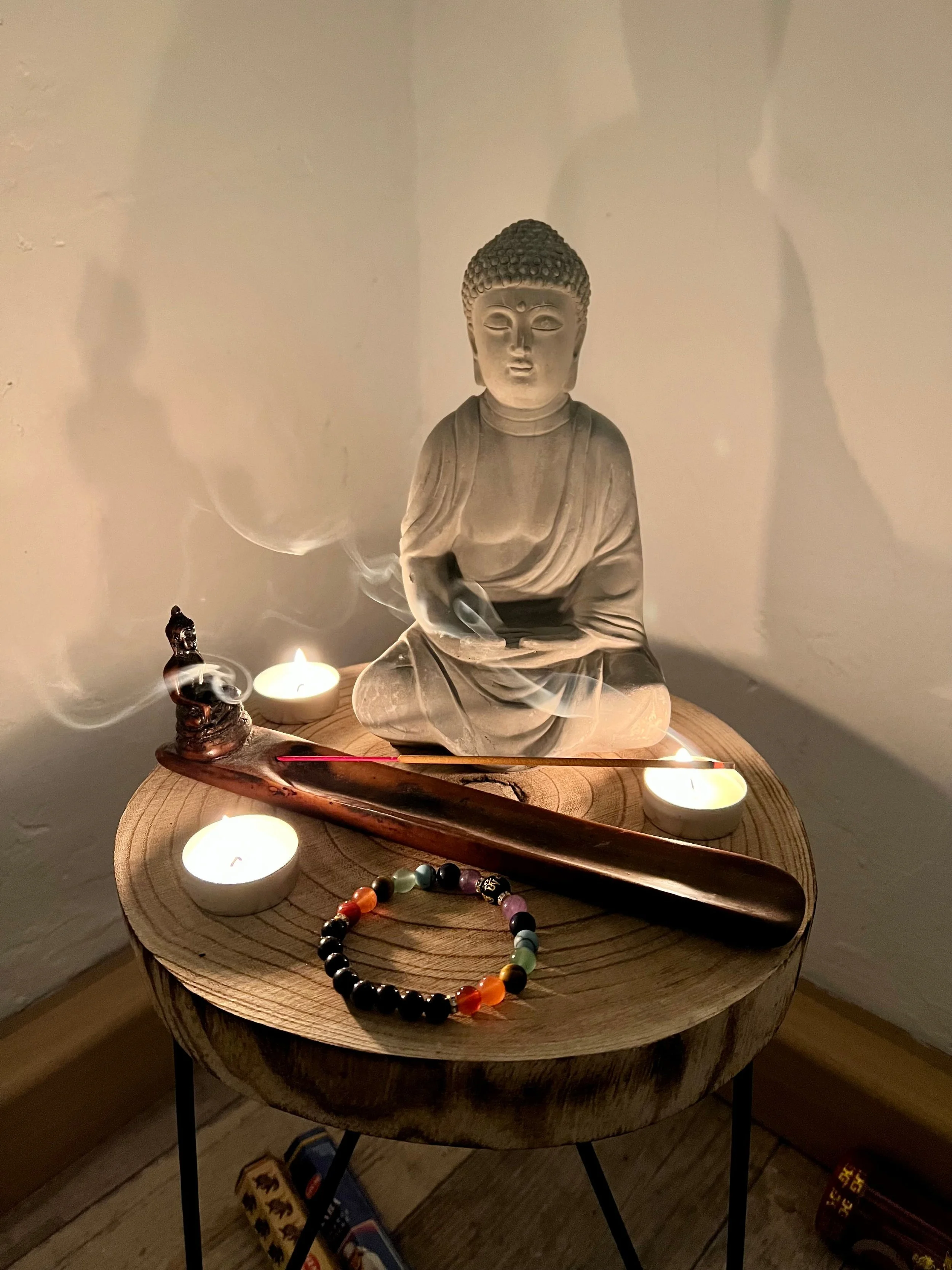 A small wooden table with a Buddha statue surrounded by candles, incense, a beaded bracelet, and a wooden stick, creating a peaceful and spiritual ambiance.