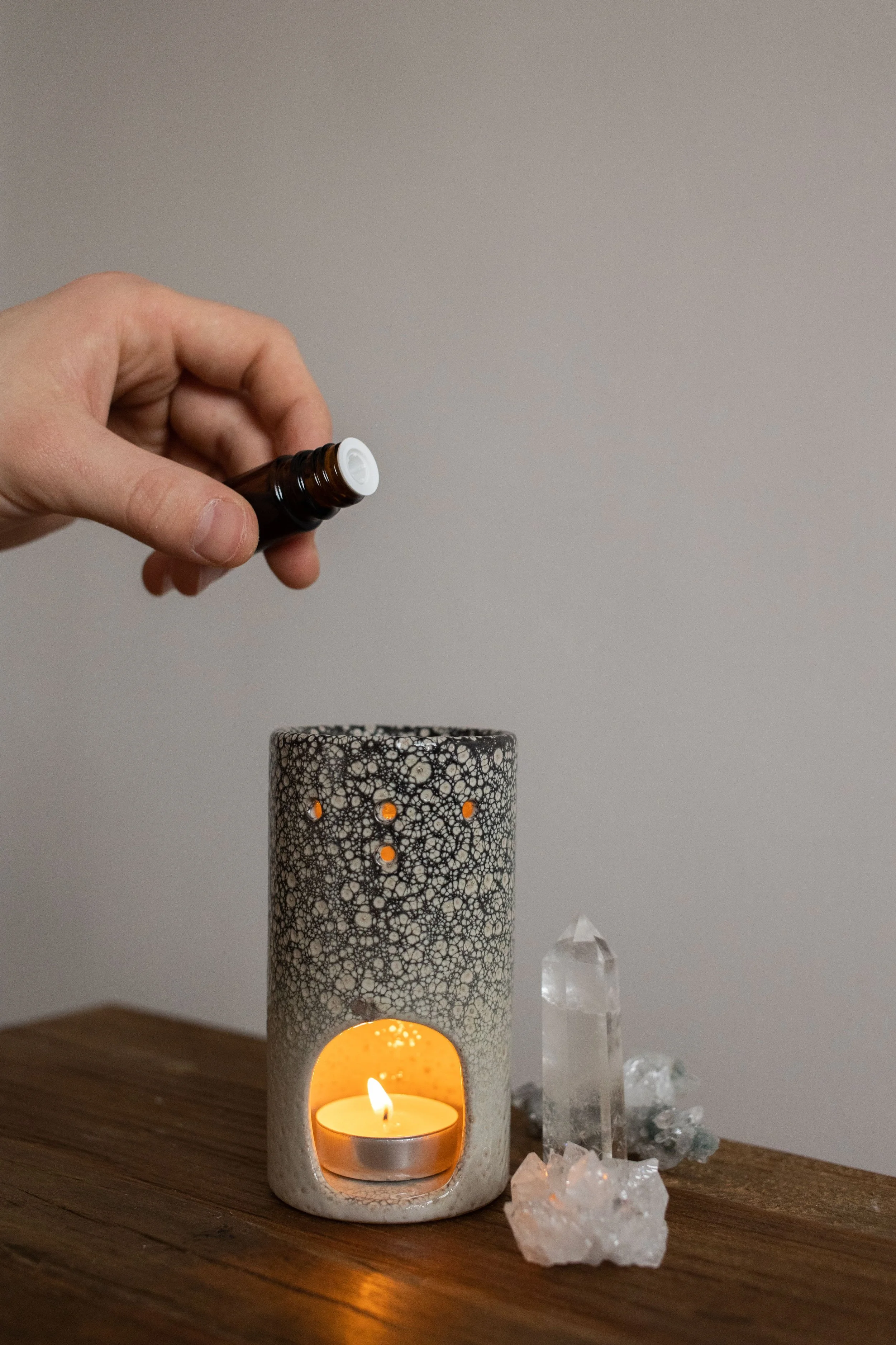 A white candle holder with intricate black lace-like design holding a lit tealight candle, surrounded by clear quartz crystals on a wooden surface, against a plain beige wall background.