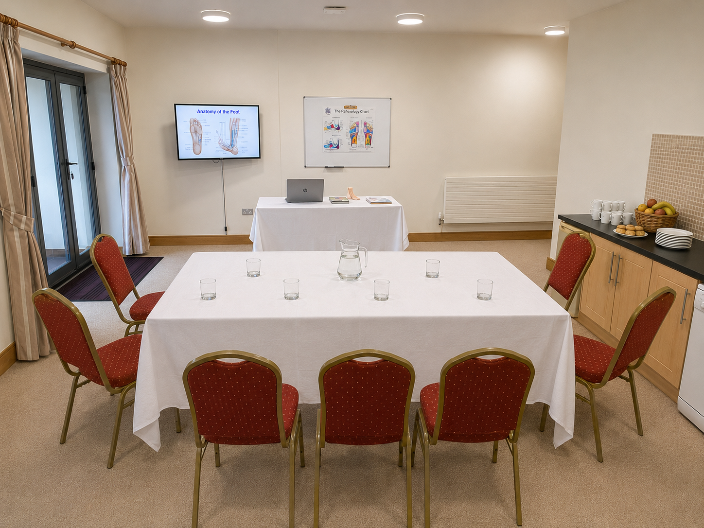 Meeting room with six red chairs around a white table, six glasses, a glass pitcher, and presentation materials on a table at the front, including a laptop and educational posters.