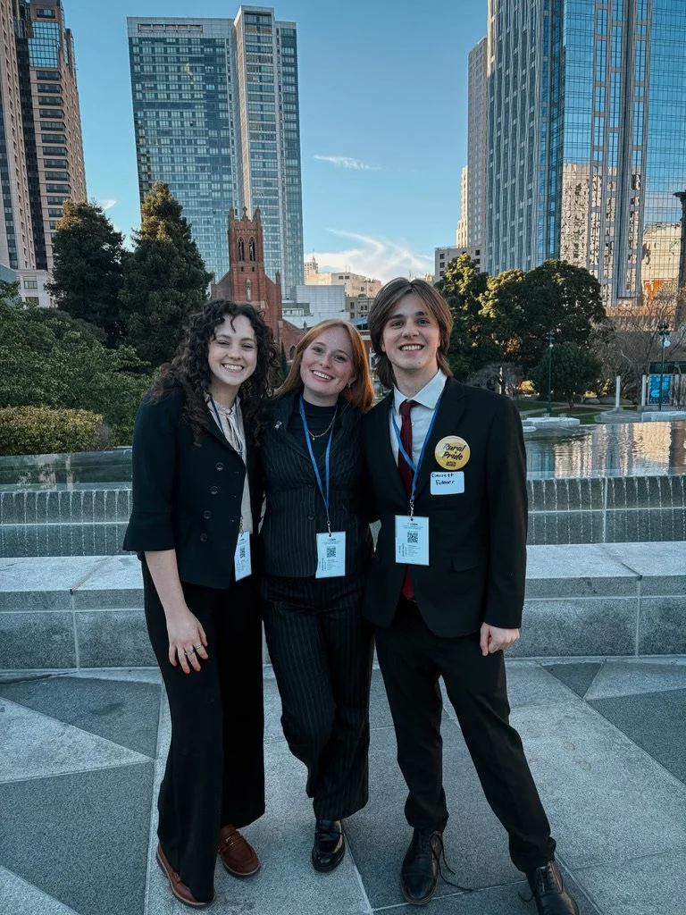 Three young women standing together outdoors in an urban park with modern high-rise buildings in the background, smiling at the camera.