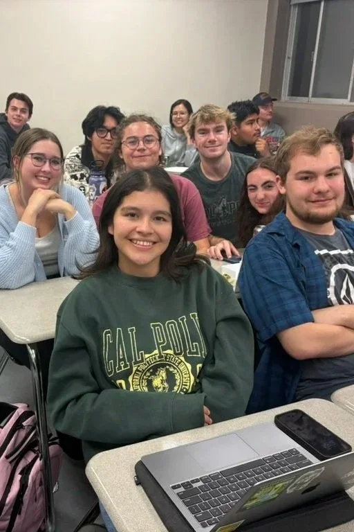 A classroom filled with diverse students sitting at desks, smiling, with a girl in the front wearing a dark green CAL POLY sweatshirt, and a laptop and smartphone on her desk.