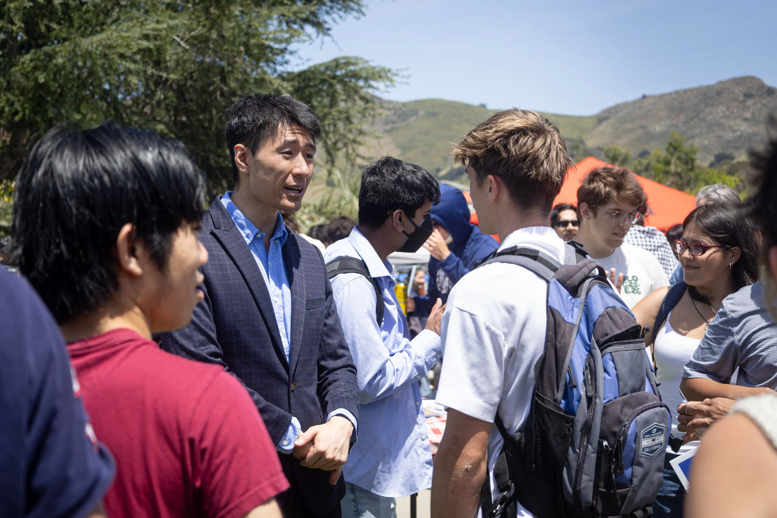 A group of diverse young people gathered outdoors, some wearing backpacks, engaged in conversation with an older man in a suit, likely at an outdoor event or tour in a scenic mountain area.