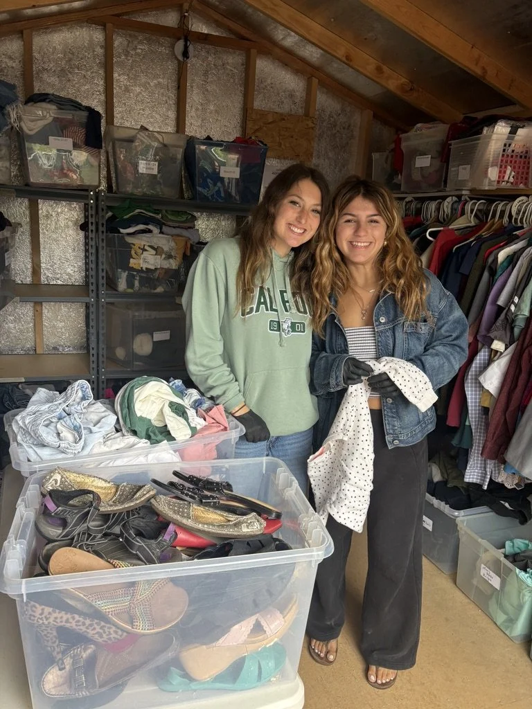 Two women smiling and holding clothes in a garage or storage room with shelves of bins and hanging clothes. One has wavy hair and is wearing a striped shirt with a denim jacket; the other has straight hair and is wearing a pastel green sweatshirt. There are shoes in a large clear plastic bin in the foreground.