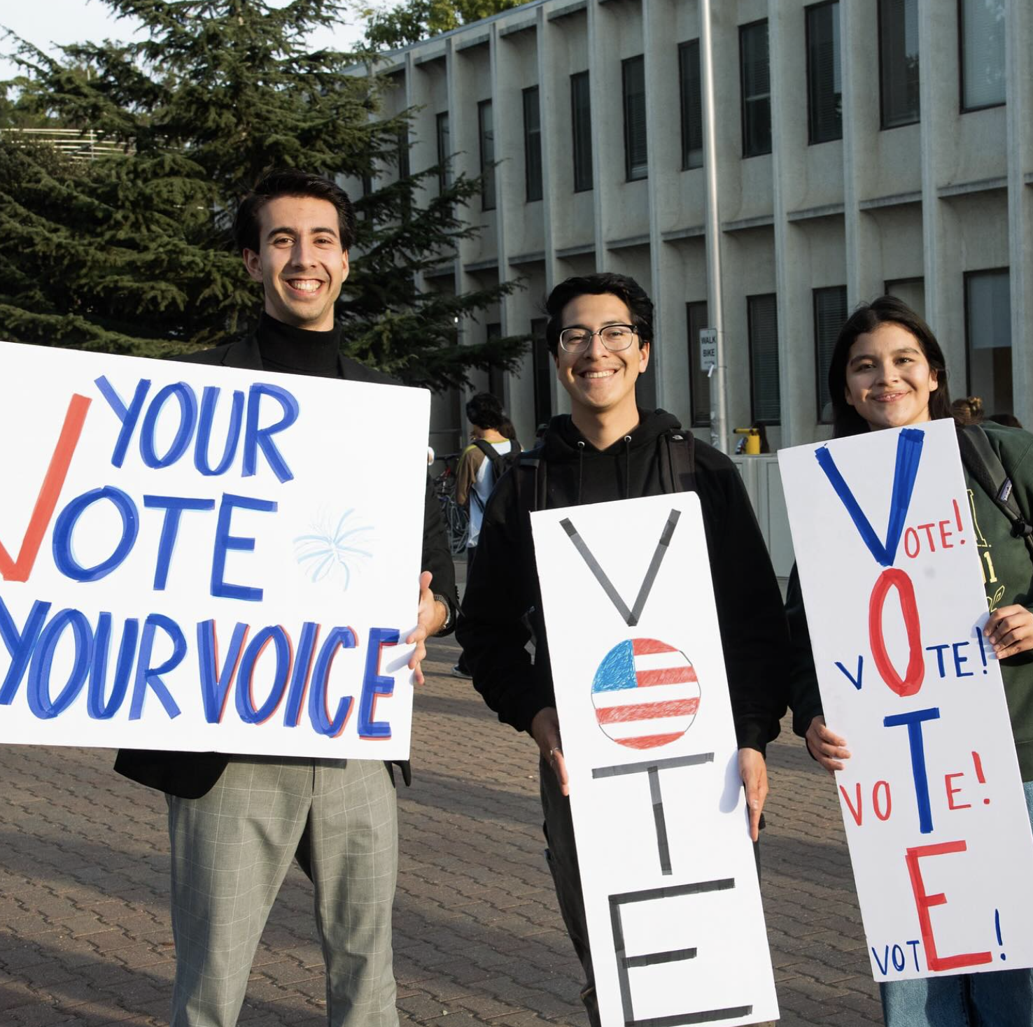 Three young adults standing outdoors holding signs promoting voting. The man on the left is smiling and holding a sign that says "Your Vote Your Voice," the man in the middle is smiling and holding a sign with a ballot box and American flag design, and the woman on the right is smiling and holding a sign that says "Vote!" with the V and T letters highlighted in red and blue. There are trees and a building in the background.