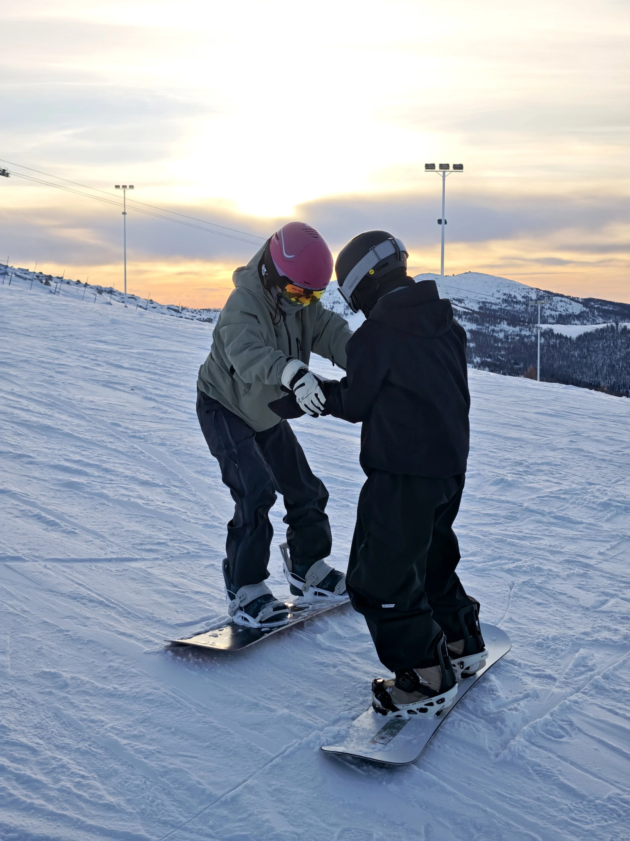 Two people, wearing ski helmets and winter gear, are on a snowy ski slope learning to snowboard together during sunset.