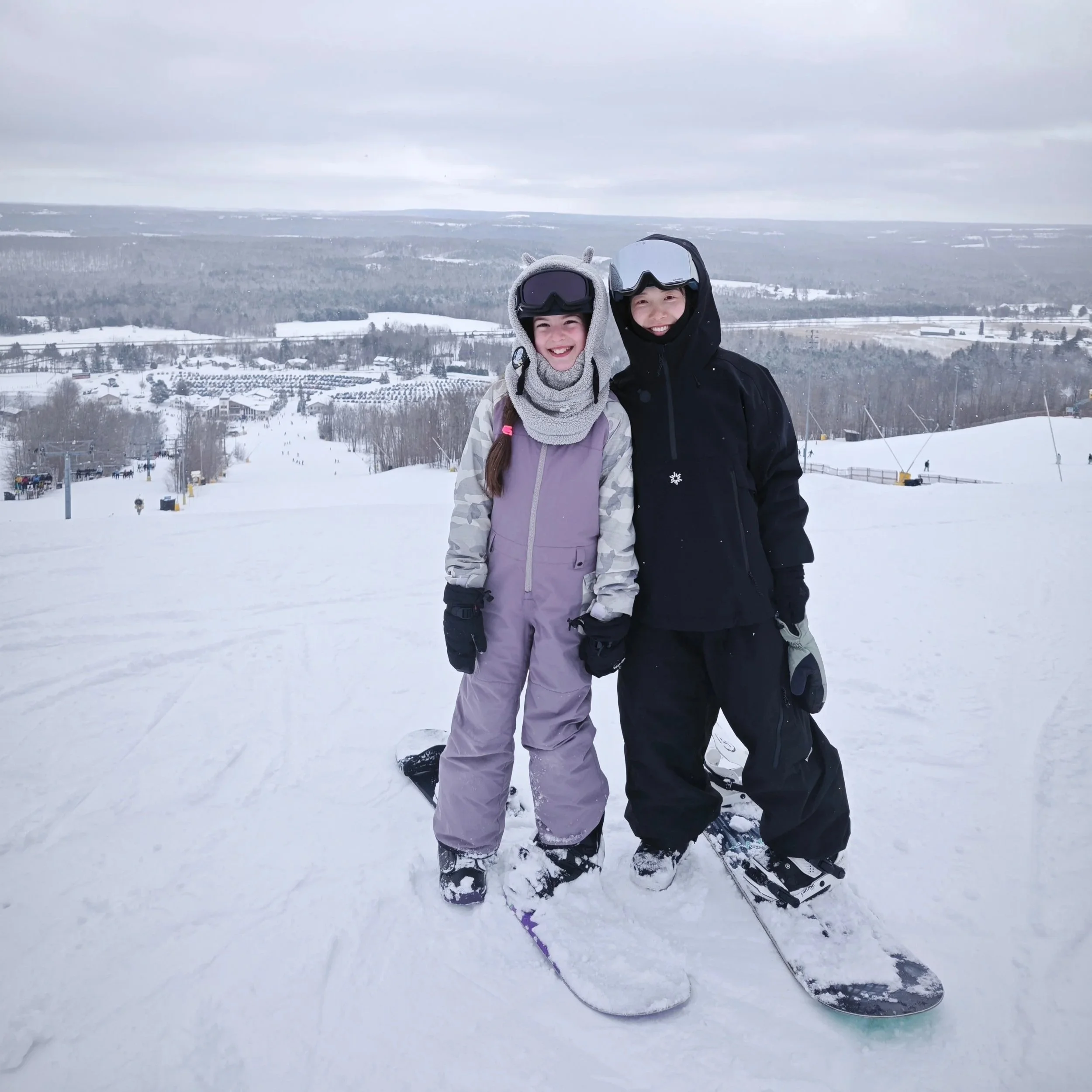 Two children in winter clothing and ski gear smiling and posing for a photo on a snowy mountain slope, with ski lifts and snow-covered landscape in the background.