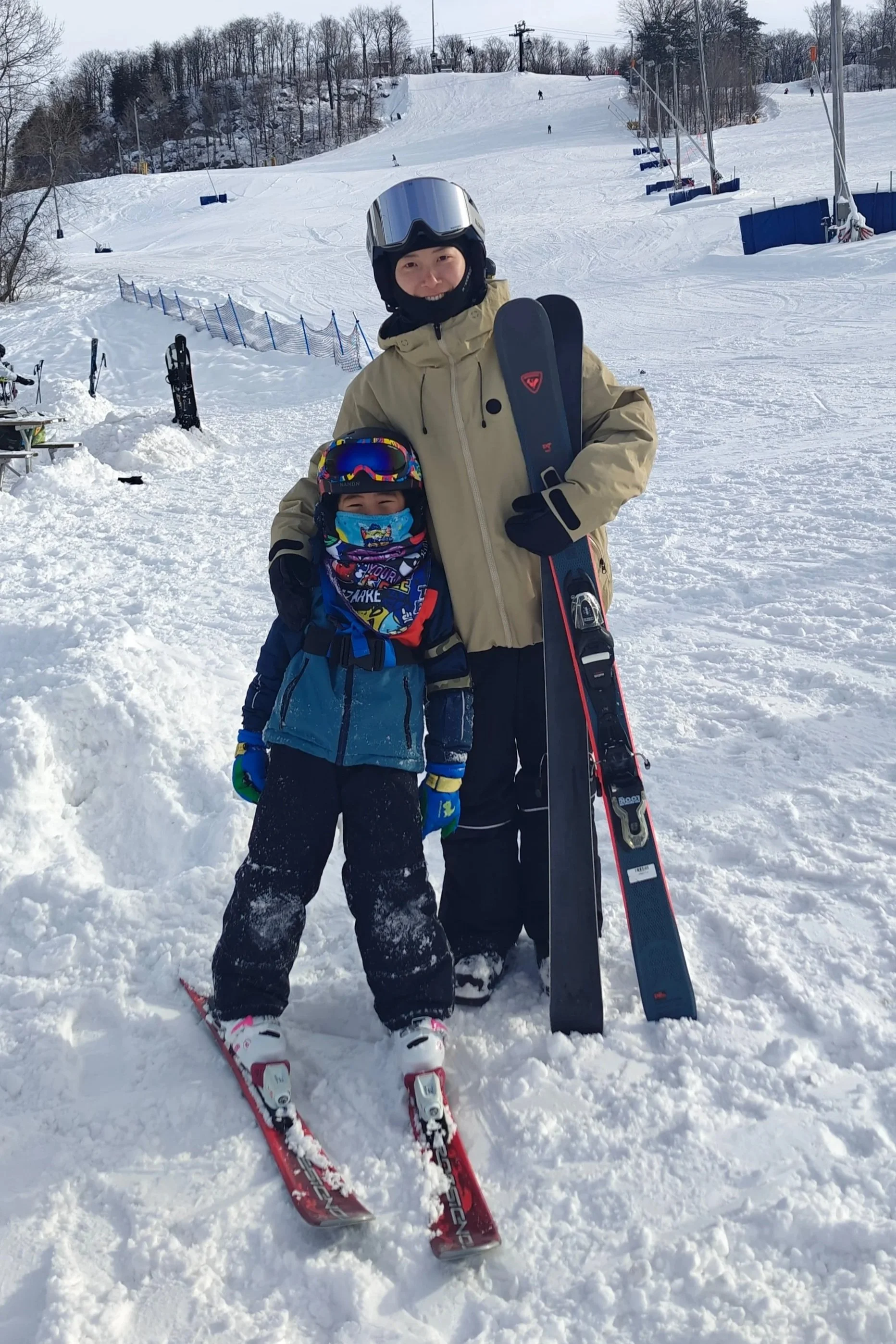A woman and a young boy standing on snowy ski slope, smiling, with skis and ski poles. The woman is dressed in beige ski gear with a helmet and goggles, and the boy in blue ski gear with a colorful face mask and helmet. Snow-covered trees and ski lif