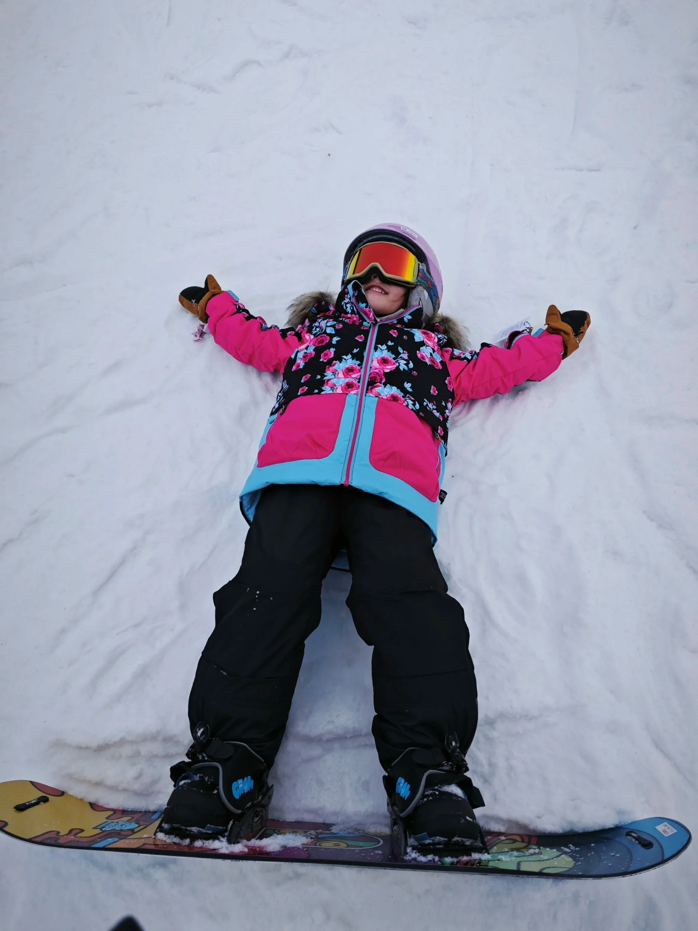 A young girl lying on snow with arms outstretched, wearing a pink and black floral winter coat, brown gloves, an orange helmet with goggles, black ski pants, and ski boots, with skis on her feet.