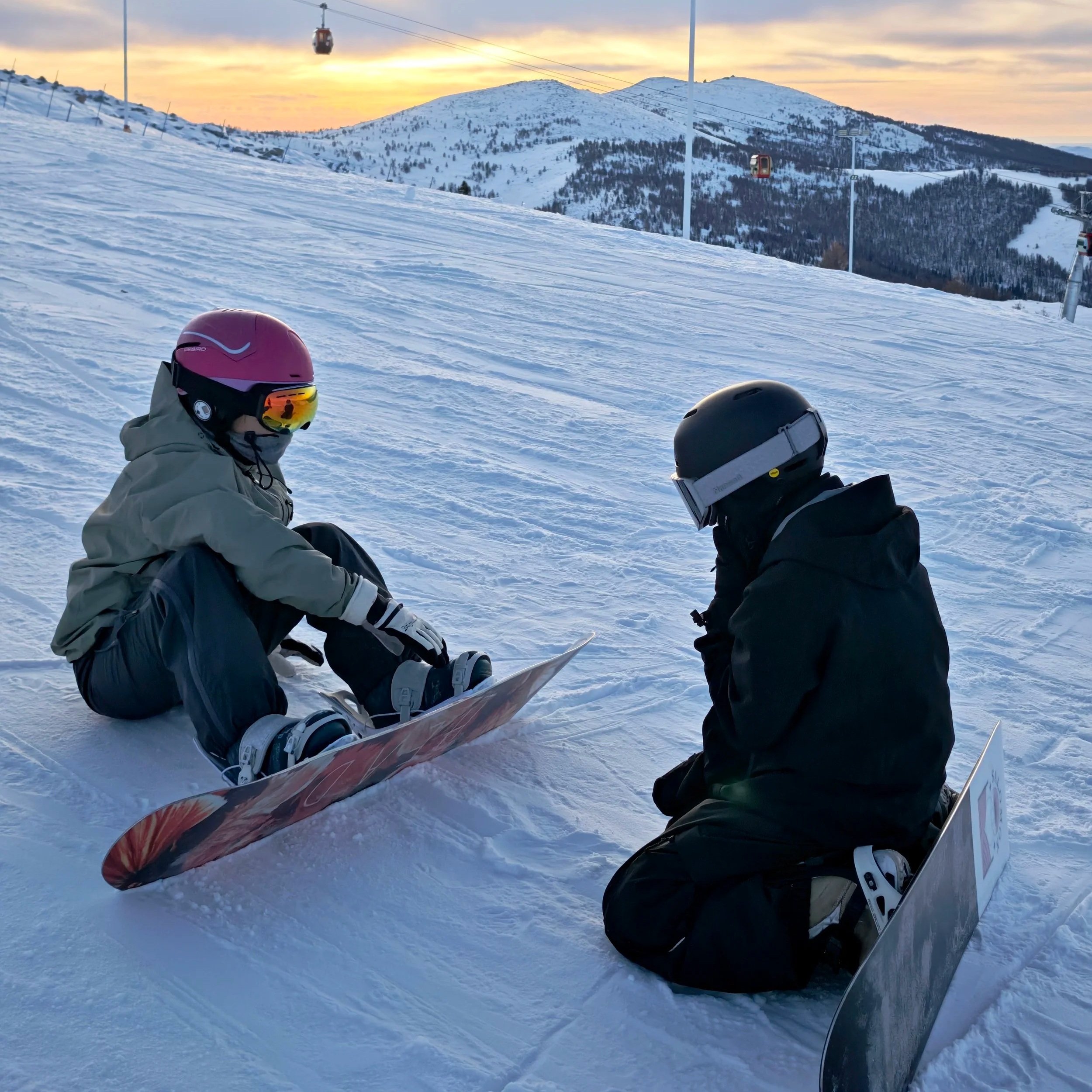People in ski gear sitting on snow at a ski resort during sunset, with ski lifts and mountain landscape in the background.