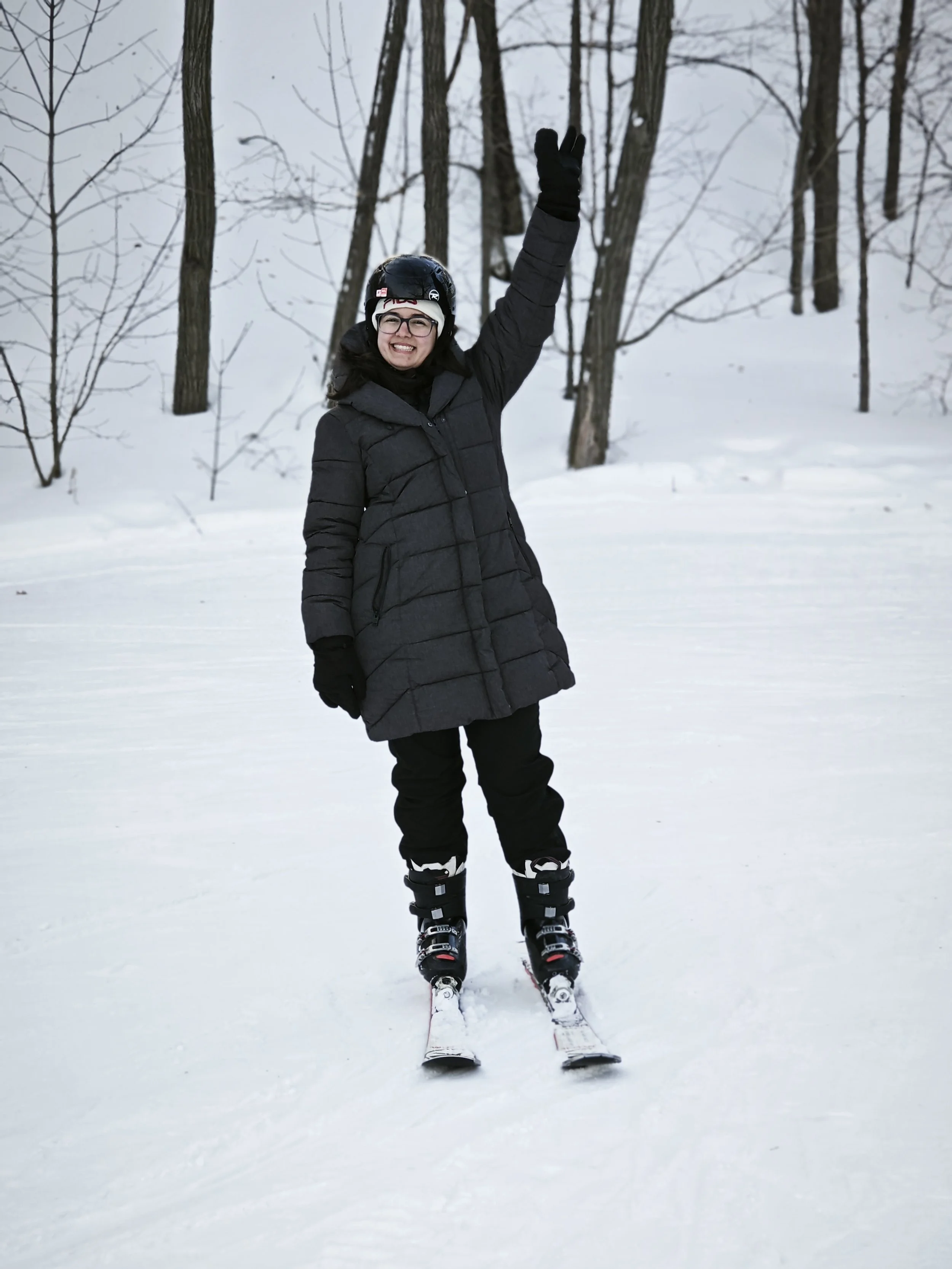A woman in winter clothing, wearing a helmet and glasses, smiling and raising her right hand while standing on skis in a snowy forest.