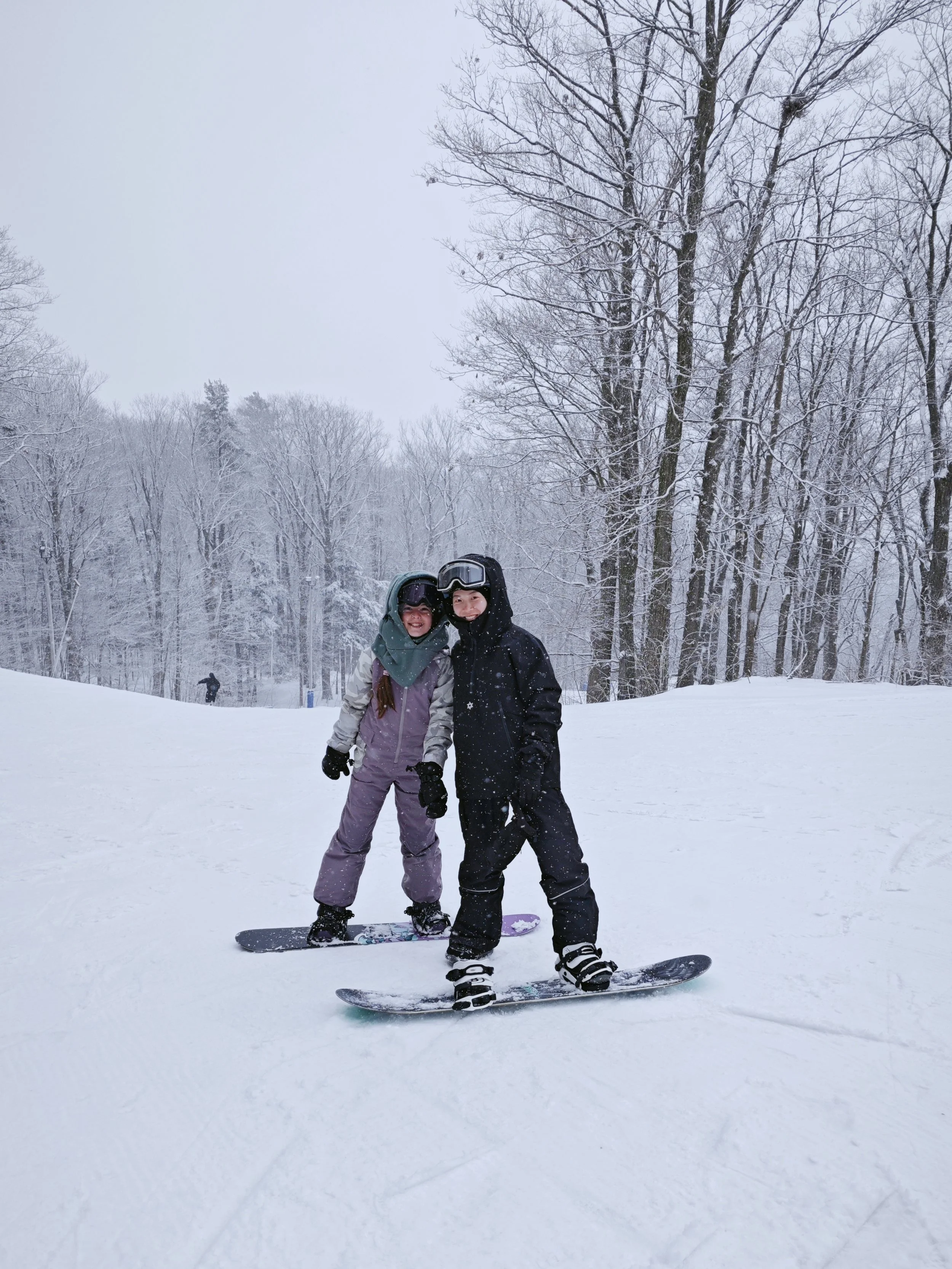 Two people in winter clothing with helmets and ski goggles on snowboards, smiling on a snowy ski slope with snow-covered trees in the background.