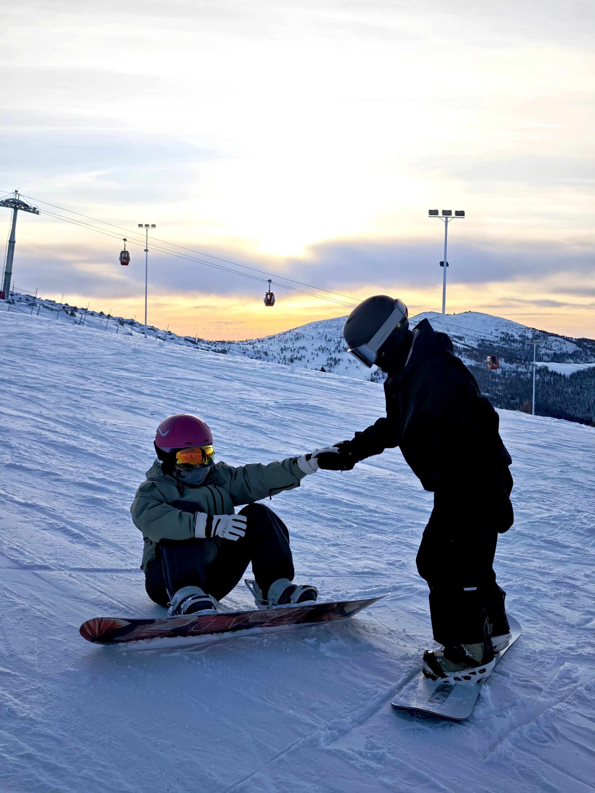 Two snowboarders on a snow-covered slope during sunset, with ski lifts and mountain scenery in the background. One person is kneeling on the snow, wearing a pink helmet and goggles, while the other is standing, wearing a helmet and snowboarding gear.
