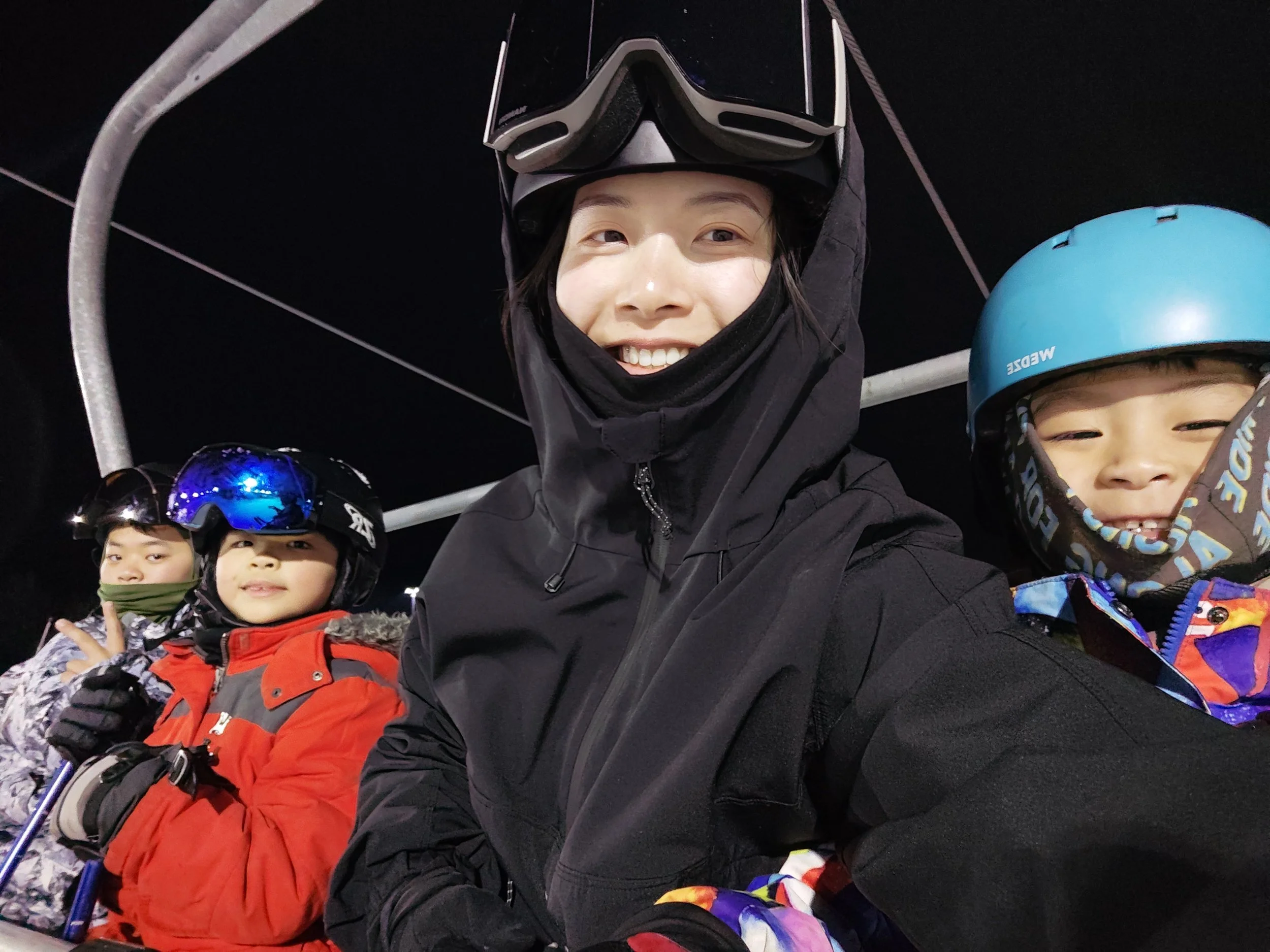 A group of smiling people in winter gear, including ski helmets and goggles, sitting on a ski lift at night.