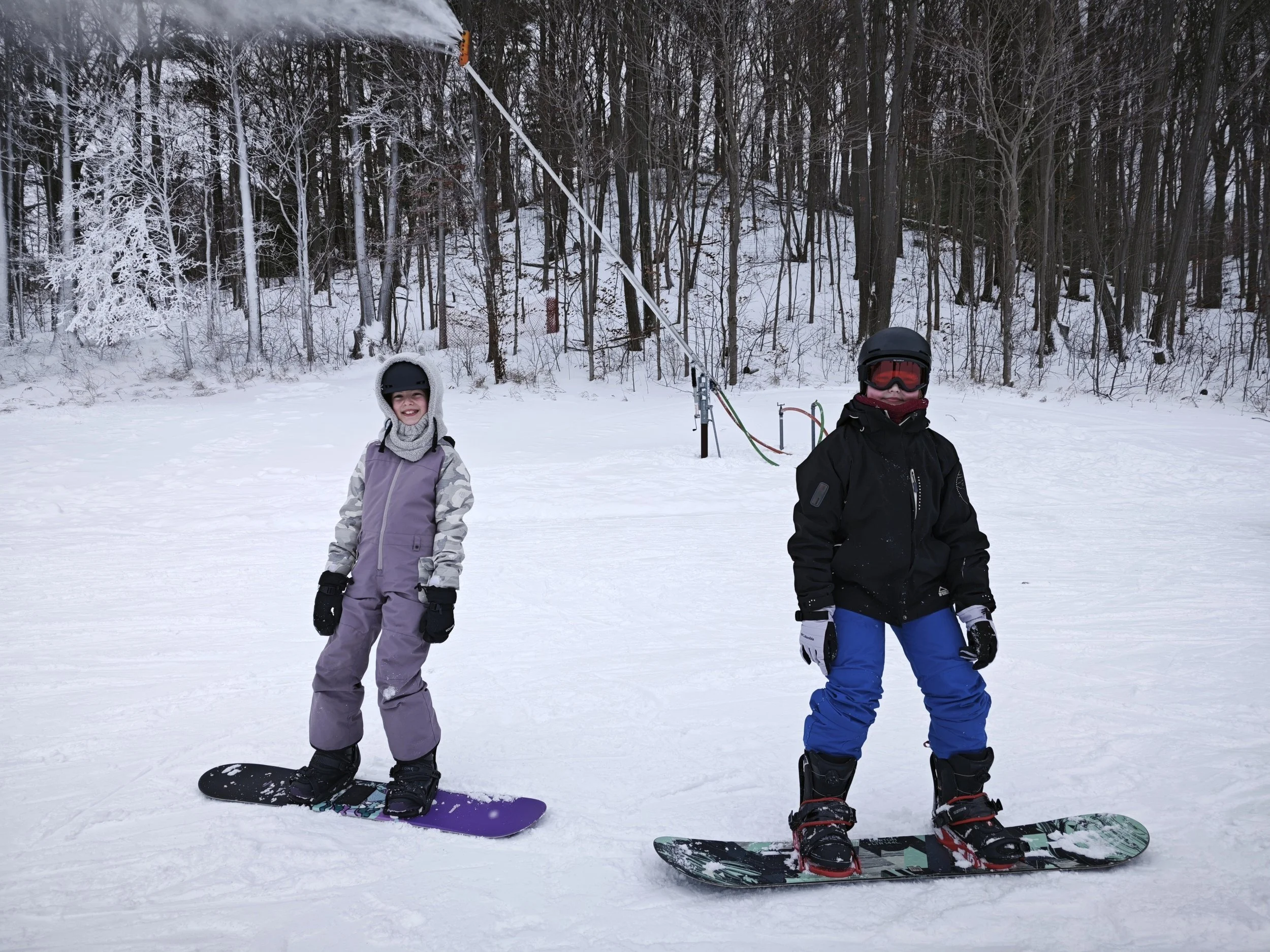 Two children on snowboards standing on snow, with a snowy forest in the background.