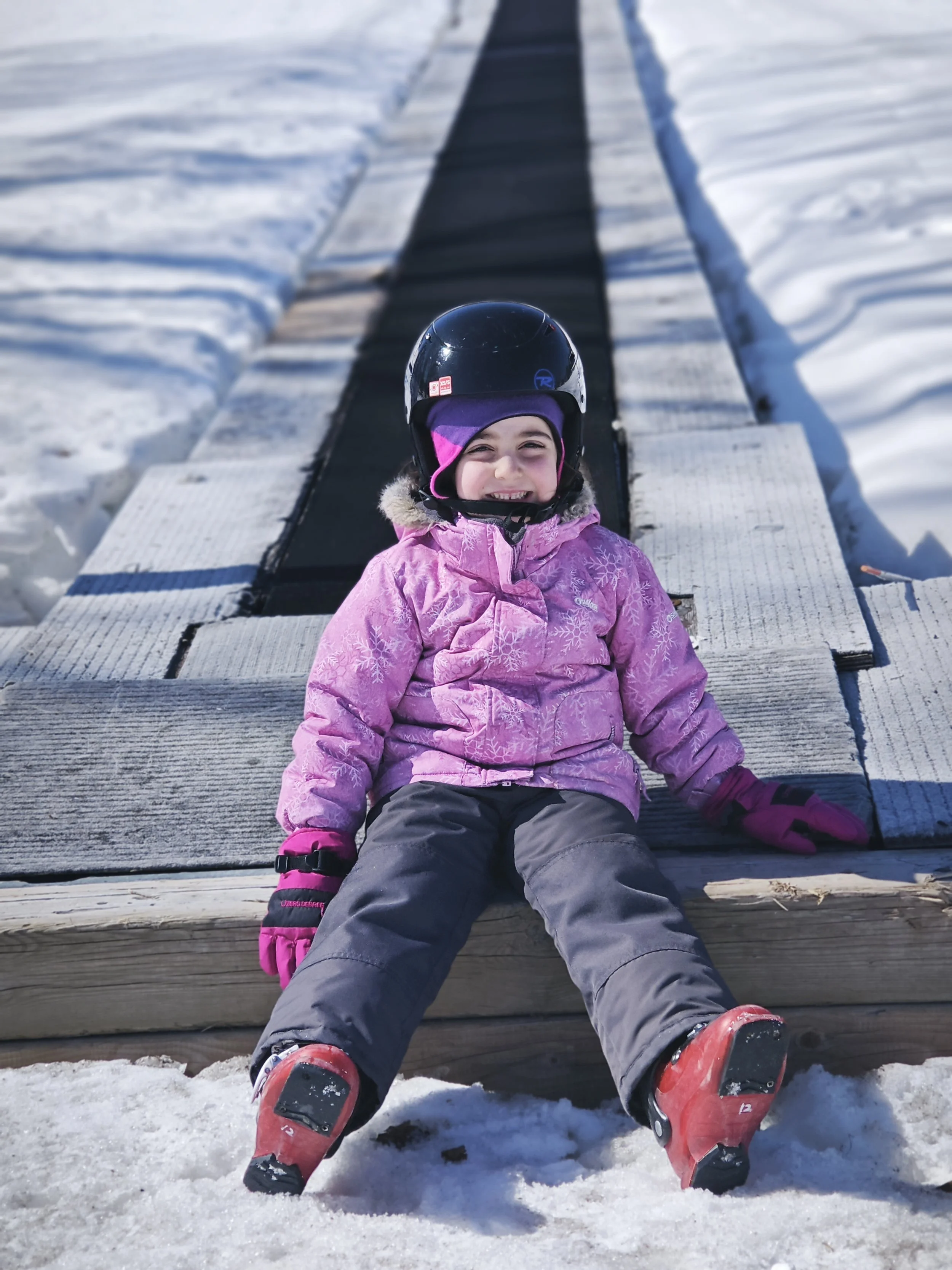 A smiling young girl in pink winter jacket, purple hat, and ski gear sitting on a wooden ramp on snow with ski boots on her feet, in a snowy outdoor setting.