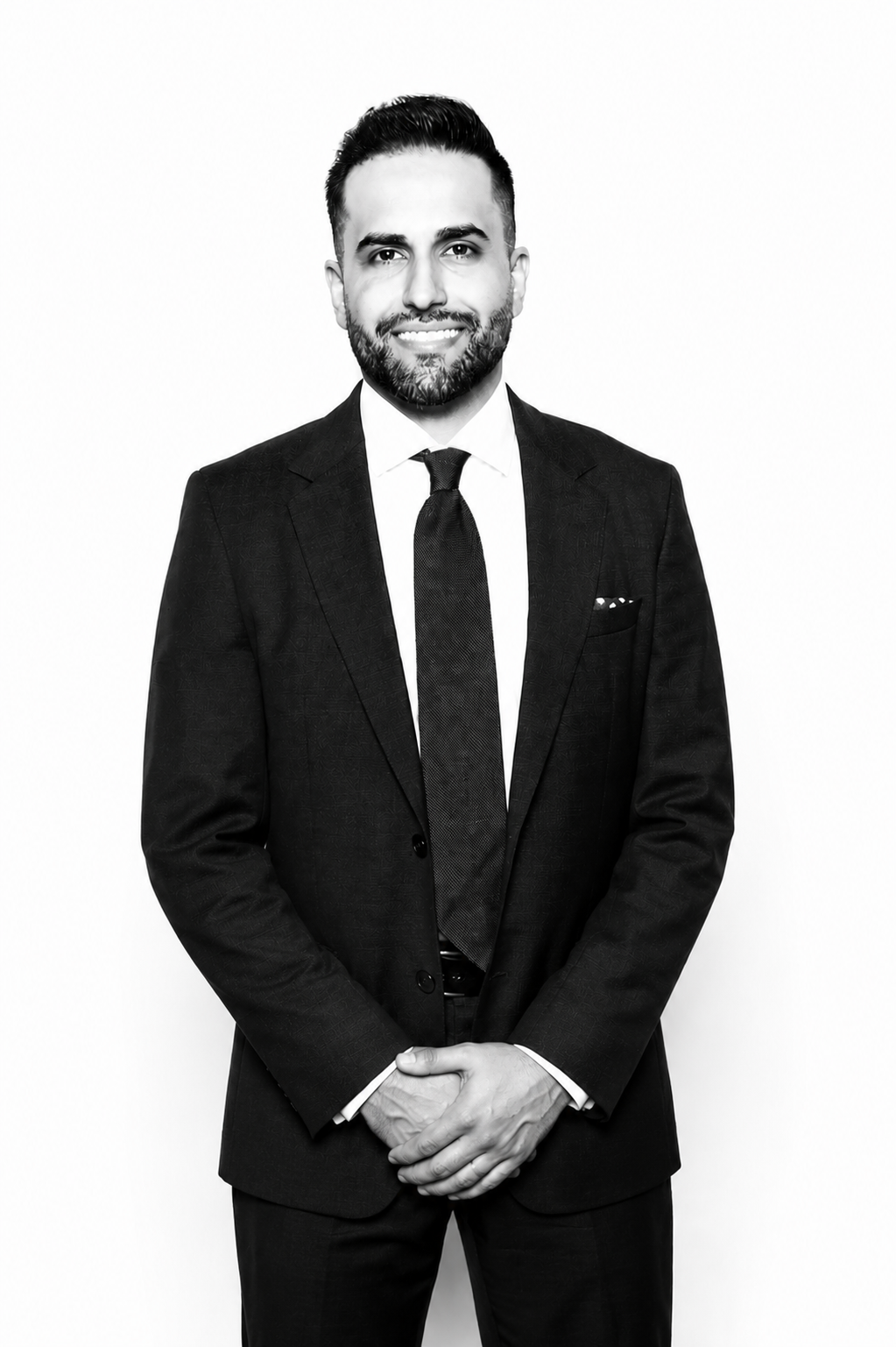 A black and white portrait of a man in a business suit, smiling, with short dark hair and a beard, standing against a plain white background.