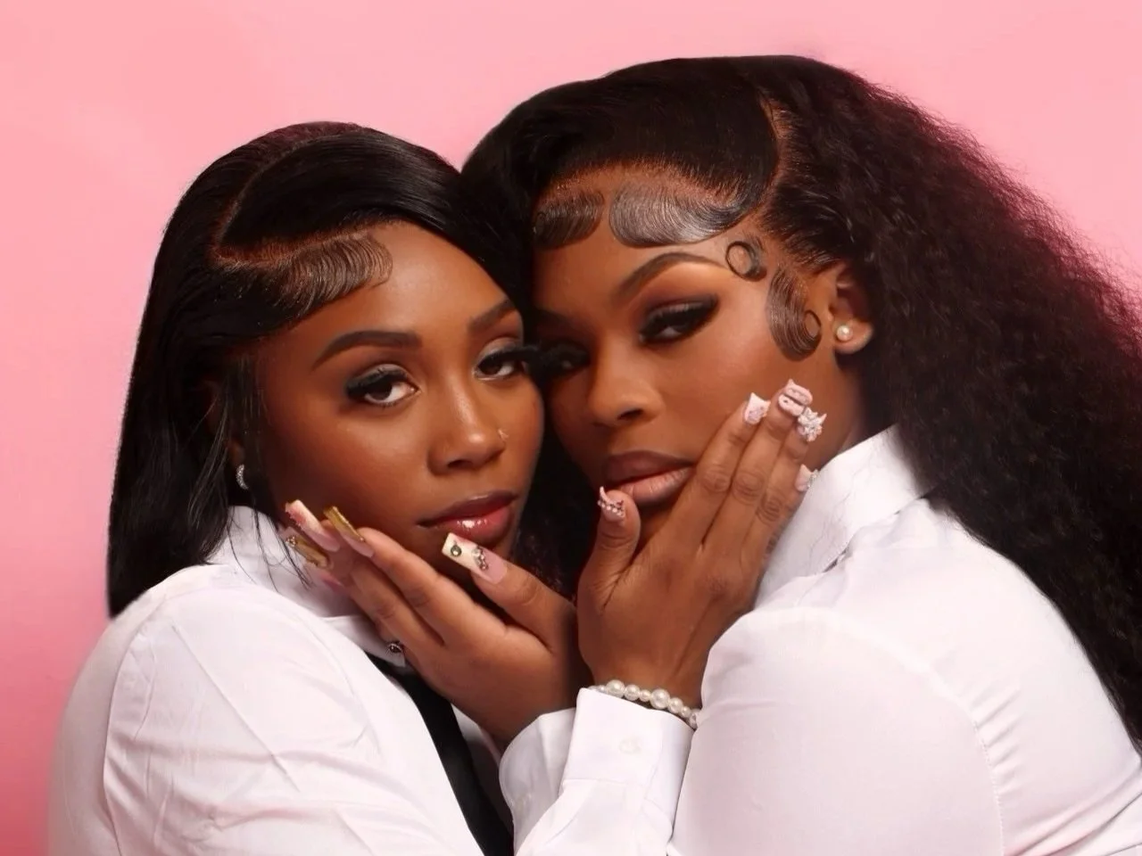 Two women with dark hair and makeup, wearing white shirts, posing closely together with hands touching their faces, against a pink background.