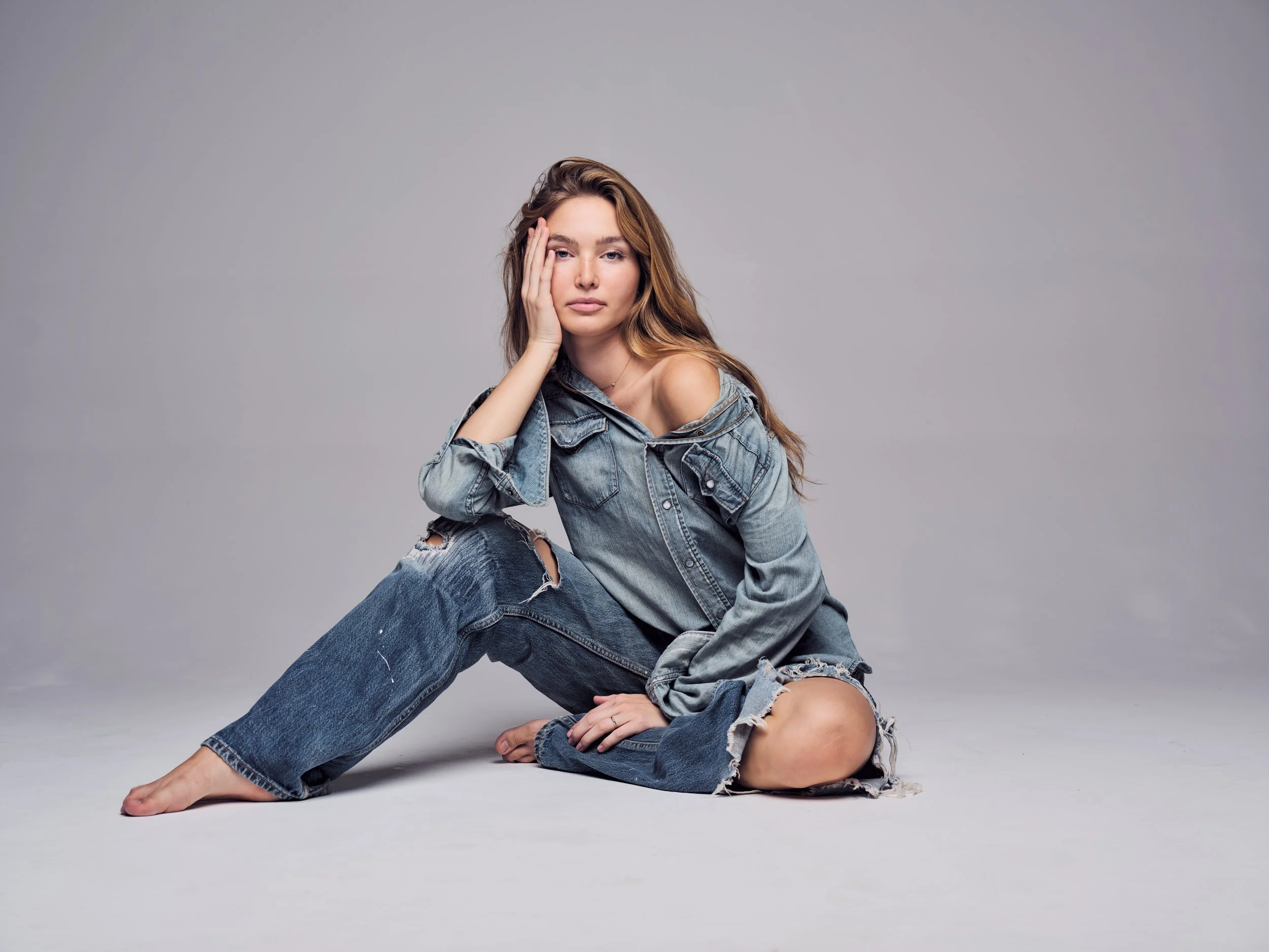 A woman with long wavy brown hair sitting on the floor against a gray background. She is wearing a distressed denim jacket and jeans, with one hand on her face and the other resting on the floor. She appears to be in a relaxed pose with a calm expres
