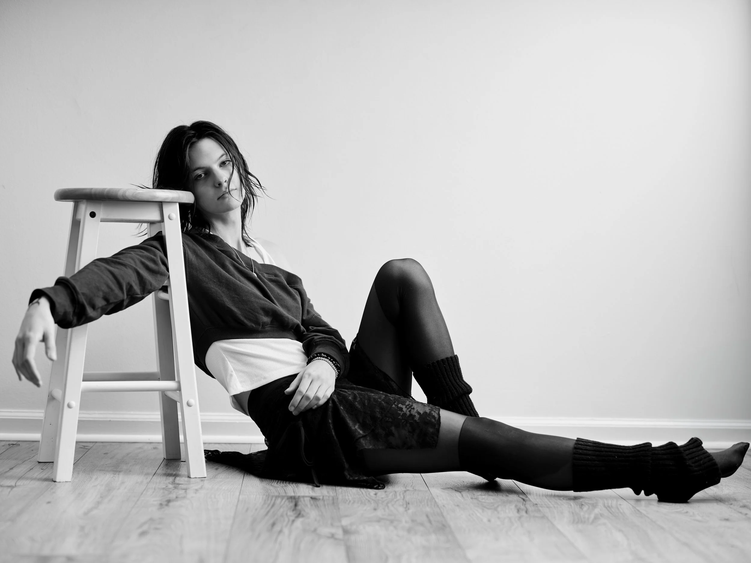 Black and white photo of a young woman sitting on the floor with her back against a wall, leaning on a chair, with her legs extended and crossed at the ankles, looking at the camera with a relaxed expression.