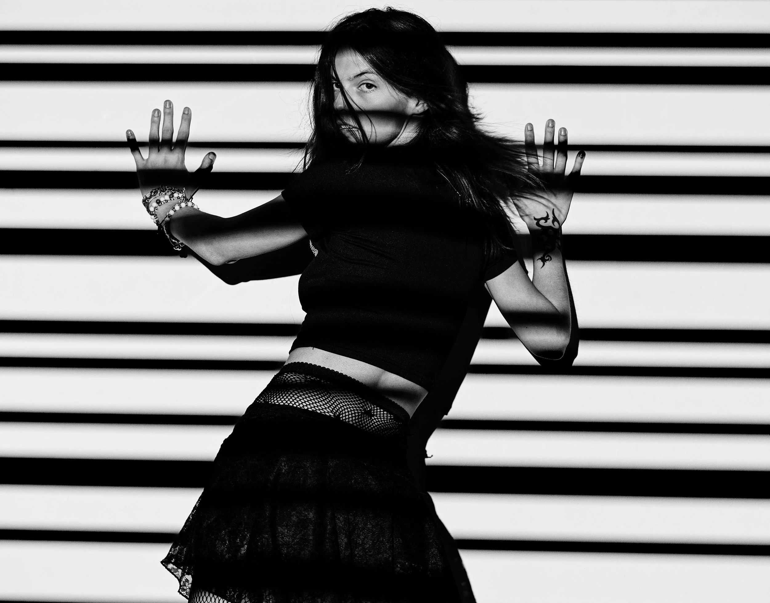 A woman with long dark hair and tattoos on her arm posing behind horizontal black and white stripes. She is wearing fishnet stockings and a dark top, with accessories on her wrist. The photo is in black and white.