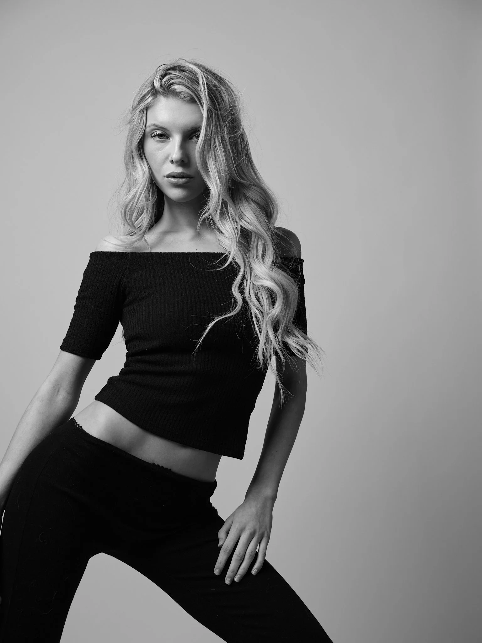Black and white photo of a woman with long, wavy hair wearing an off-the-shoulder top and black pants, posing against a plain background.