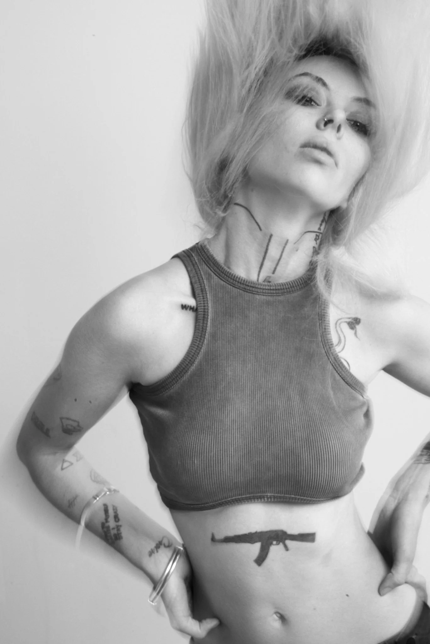 Black and white photo of a woman with tattoos on her arms and a gun tattooed on her waist, wearing a sleeveless shirt, with messy hair, and makeup, looking at the camera.