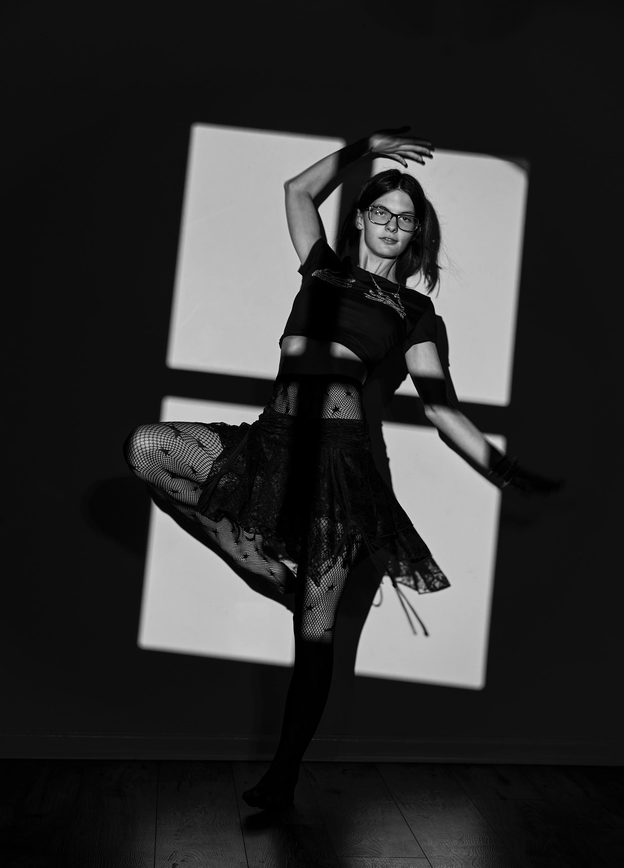 A woman with glasses and dark hair, dressed in a black top, fishnet tights, and a tulle skirt, poses with her right arm raised above her head and her left leg bent at the knee. She is illuminated by a rectangular light source behind her, creating a h