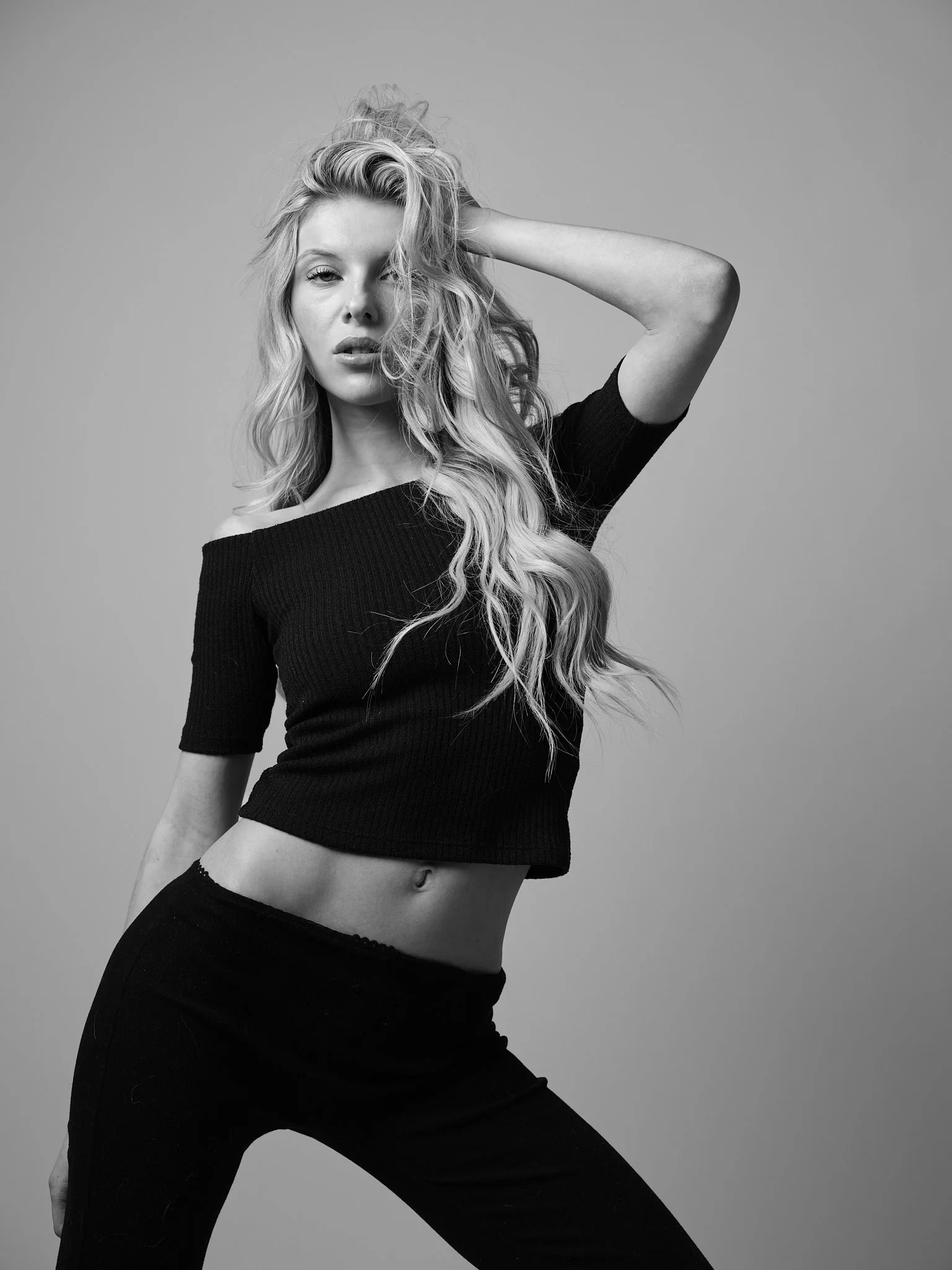 Black and white photo of a woman with long, wavy hair, wearing a off-shoulder top and black pants, posing with one arm raised behind her head.