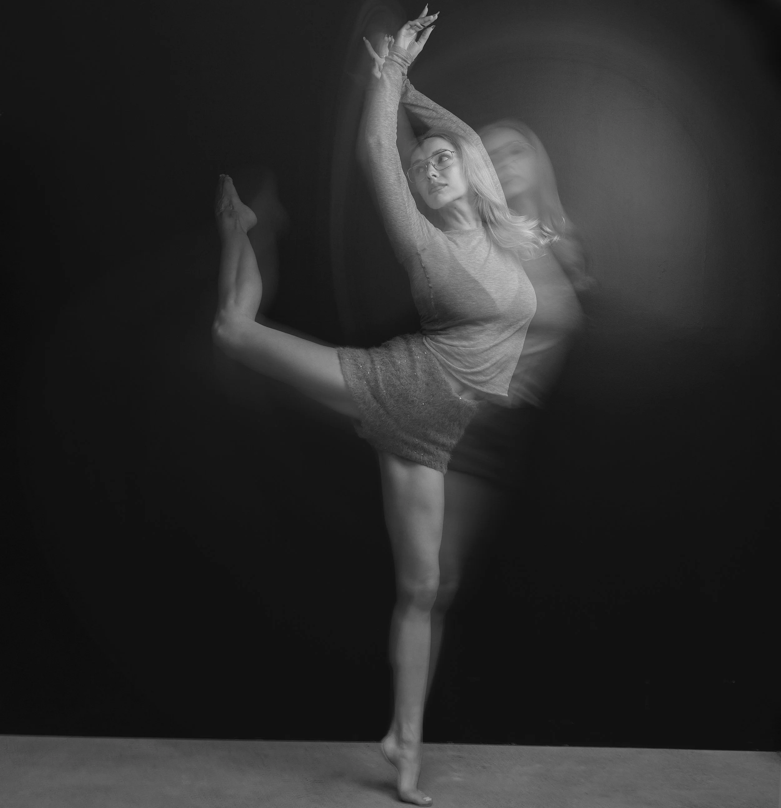 Black and white photo of a woman performing a dance pose, balancing on one foot. She has long hair, glasses, and is wearing a long-sleeve top and skirt. Her arms are raised above her head, and her expression is focused. The background is plain and da