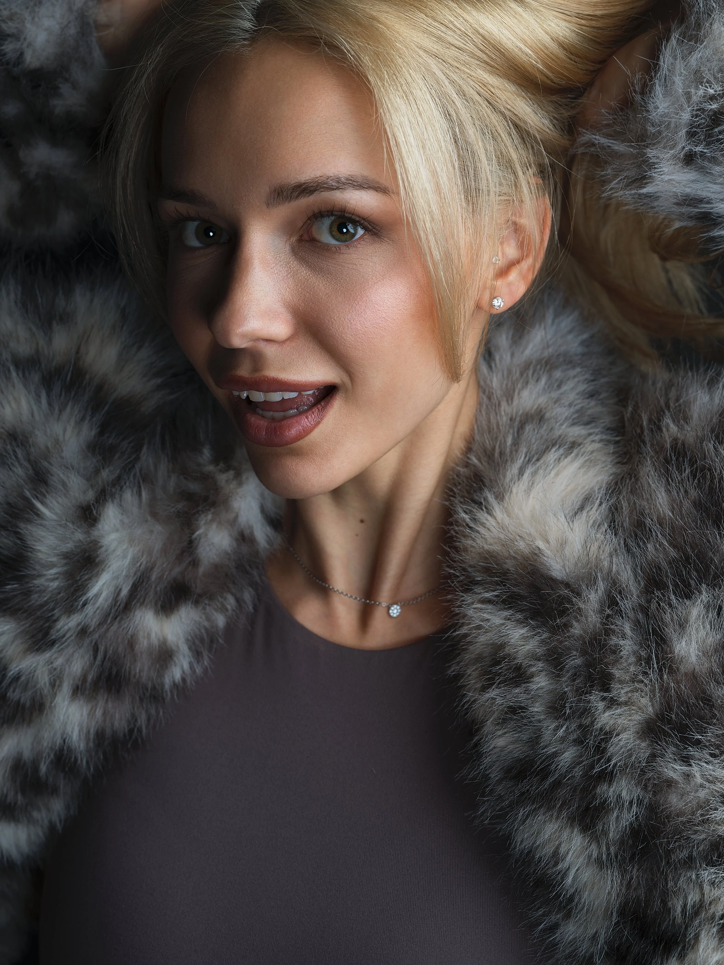Close-up portrait of a woman with blonde hair, green eyes, and diamond earrings, wearing a gray top and a fur coat, with a surprised expression.
