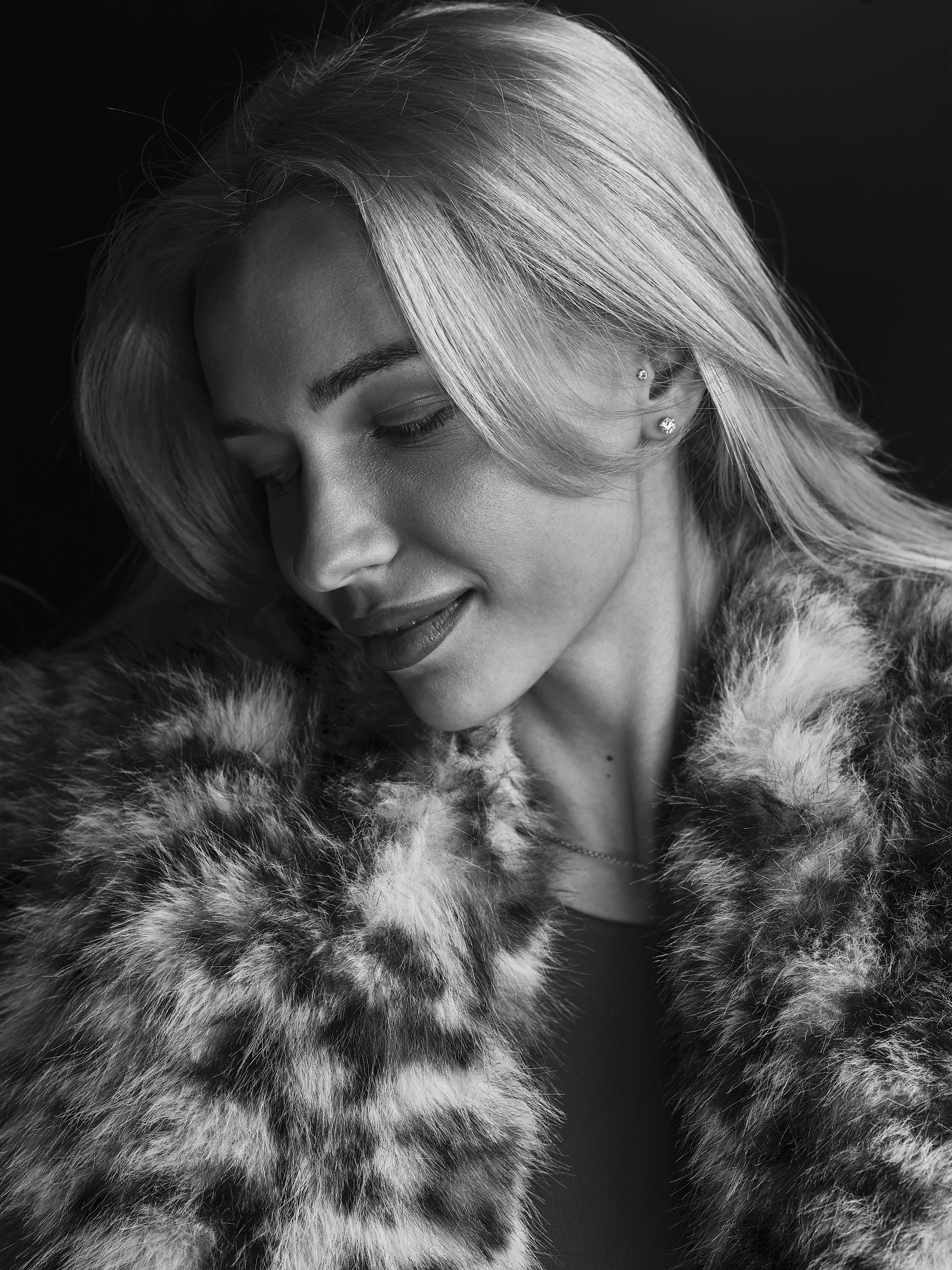 Close-up black-and-white portrait of a woman with blonde hair, wearing a fur coat, with quit earrings, her eyes closed, and a slight smile.